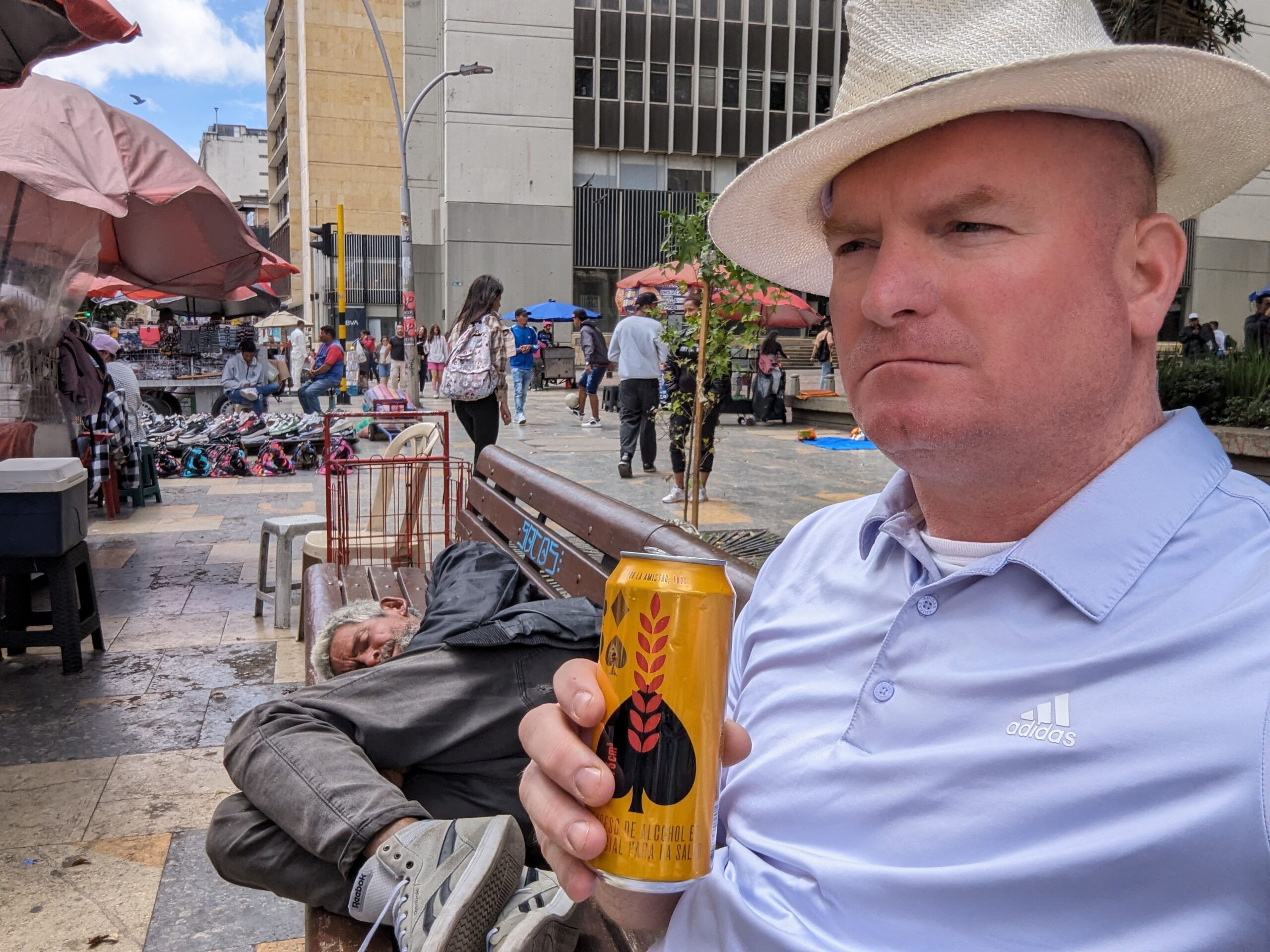 Michael McGill sits on a bench in Bogota Colombia drinking a can of Poker Beer next to a sleeping homeless man in the park.