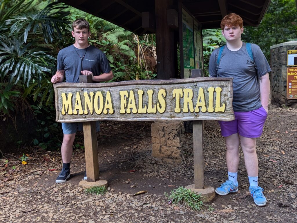 Two individuals stand beside a wooden sign that reads ‘MANOA FALLS TRAIL.’ The sign is mounted on two stone pillars and situated at the trailhead, with lush greenery in the background indicating a forested area. The individuals’ faces are obscured for privacy.