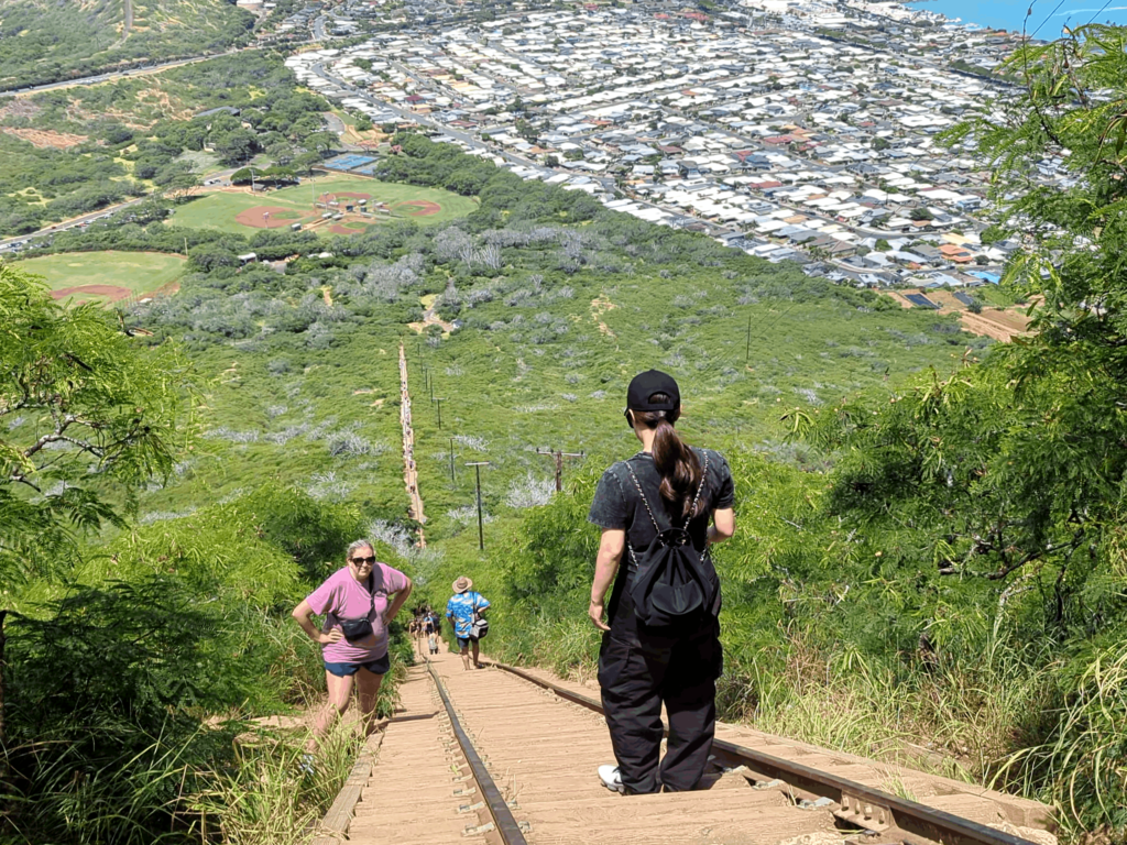 A scenic view from hiking down Koko Head trail. In the foreground, an individual stands on train tracks, facing away from the camera and looking out towards the landscape. Another person in pink attire is ascending the trail. The surrounding vegetation is lush and green, and in the distance, there’s a panoramic view of a Honolulu leading up to an expansive ocean under a clear blue sky.