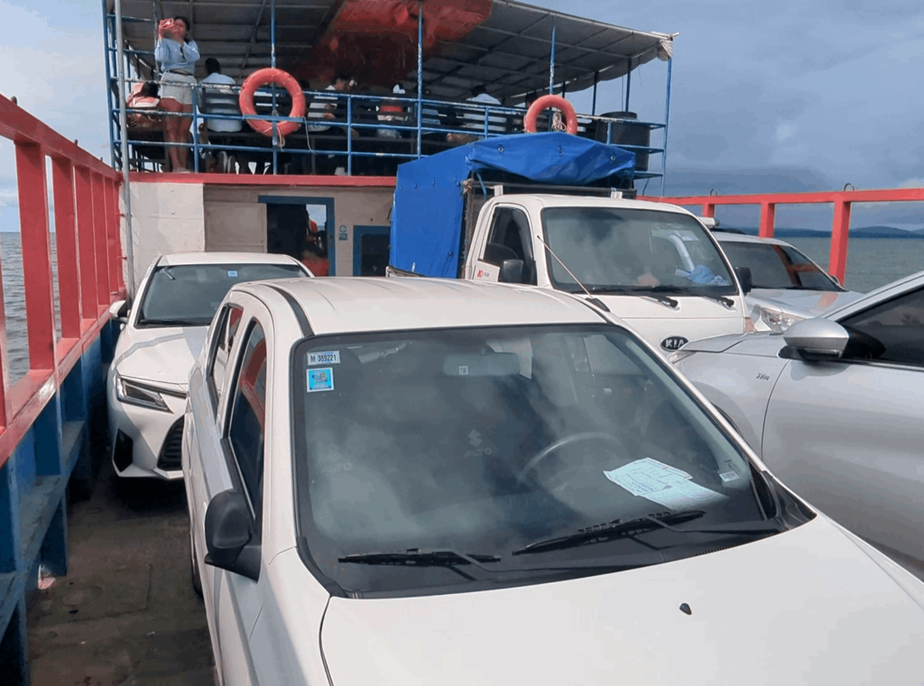 A variety of vehicles, including cars and a truck, loaded onto a ferry heading for Ometepe Island in Nicaragua. The ferry has two visible decks; the upper deck has people standing by the railing, looking out over the water. Lifebuoys are attached to the railing for safety. The sky is overcast with clouds, suggesting it might be a cool or rainy day.