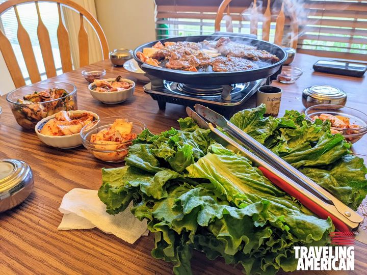 A dining table set for a meal, featuring Korean Bulgogi with a large bowl of lettuce in the foreground and a hot grill pan with cooking meat at the center. Surrounding the grill are various small dishes containing different side dishes, likely Korean banchan. The setting suggests a communal dining experience, possibly Korean BBQ, highlighting cultural cuisine and the social aspect of sharing food.