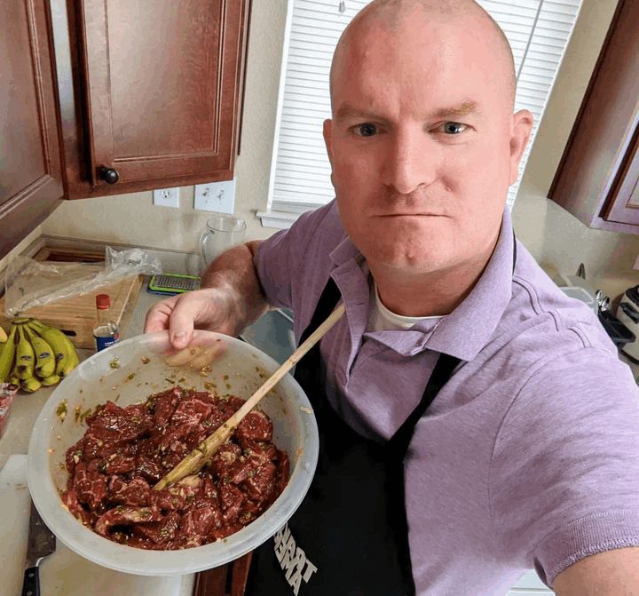 An individual in a kitchen, wearing a purple shirt and a black apron that read Traveling American, mixing a bowl of marinated Korean Bulgogi meat with a wooden spoon. The person’s face is pixelated for privacy. This scene highlights meal preparation in a home kitchen