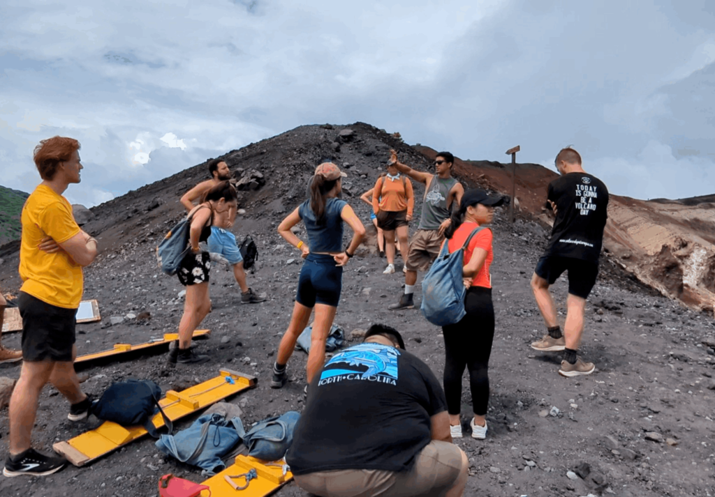 A group of individuals stands at the summit of Cerro Negro a volcanic landscape, with one person explaining the history of Cerro Negro interacting with the tour group The terrain is rocky and barren with various shades of brown and green, indicative of volcanic activity. In the background, another peak can be seen under a cloudy sky, adding to the dramatic scenery. The individuals are dressed in casual hiking attire suitable for such an environment, suggesting they are tourists or hikers exploring the area.