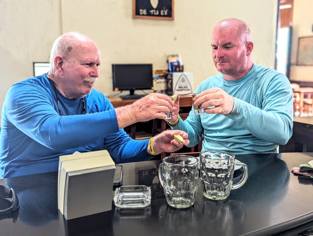 Ron and McGill are seated at the bar at ViaVia León in Nicaragua, engaging in a victory toast for sleding doing a Volcano with shot glasses. In front of them on the table is an open box, a clear empty beer mugs, and another object that resembles a lime or lemon wedge. The setting suggests a casual social gathering, at a bar called ViaVia León, indicated by the presence of drinkware and the relaxed posture of the individuals.