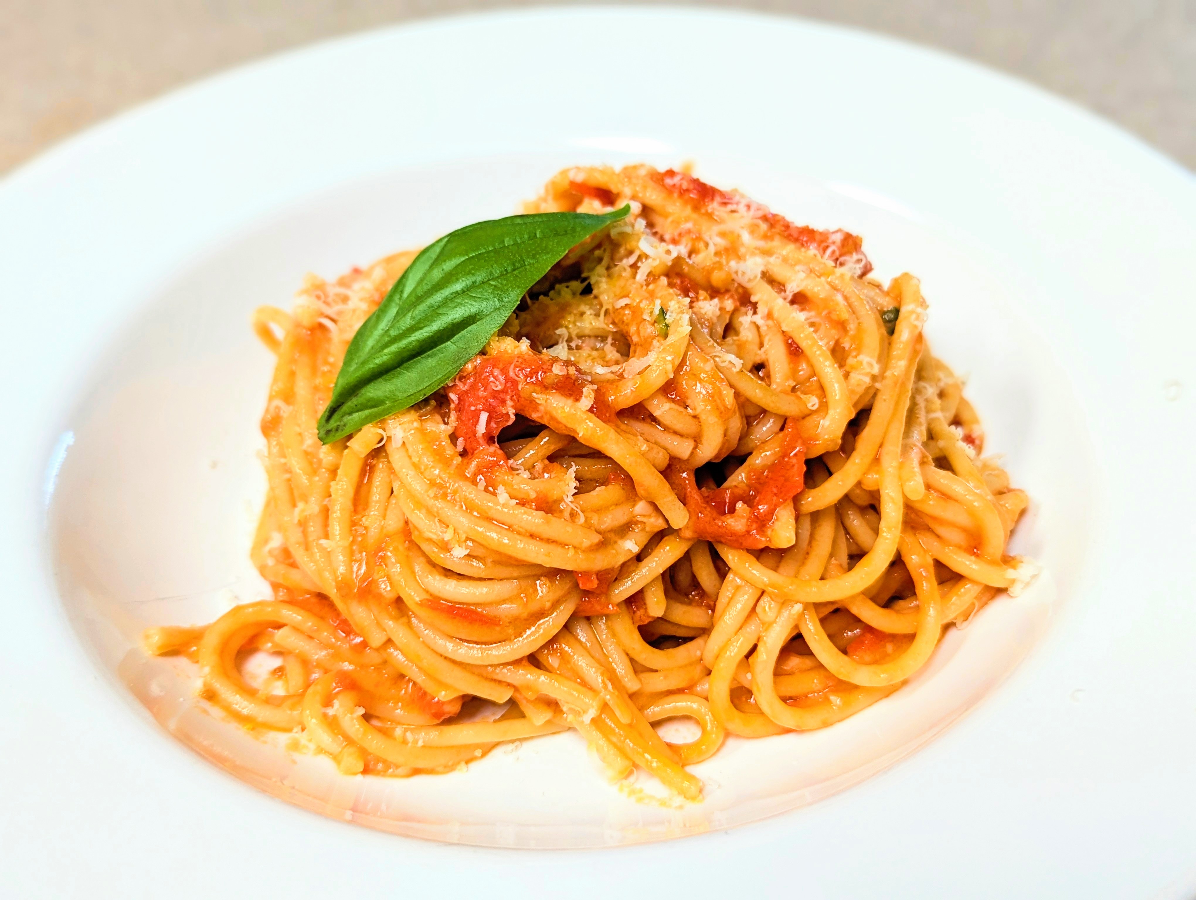 A plate of Spaghetti Pomodoro with tomato sauce garnished with a single fresh basil leaf on top. The pasta is twirled into a mound in the center of a white plate, with chunky tomato sauce and a sprinkling of grated cheese.”