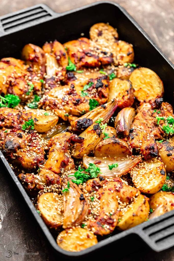 A black baking tray filled with Syrian Baharat Baked Chicken Thighs which is roasted chicken pieces and potato halves, garnished with sesame seeds and chopped parsley. The chicken and potatoes have a golden-brown crust, indicating they are well-cooked. The dish appears juicy and flavorful, suggesting a savory meal option.