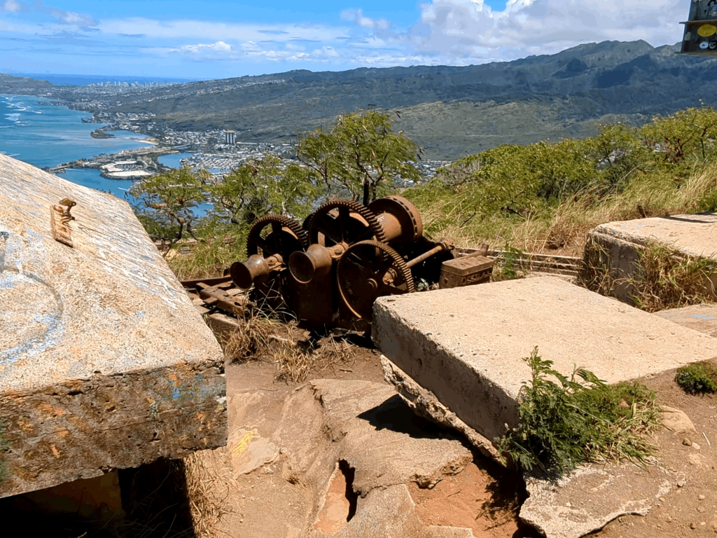 A scenic view Honolulu Hawaii from the top of Koko Head in Hawaii overlooking Honolulu with clear blue skies and some clouds. In the foreground, there is rusted wench that sit atop Koko Head that was used in WW II to pull Soldiers and Equipment to the observation post on , possibly part of an old winch or hoisting system, set on a concrete base with graffiti. The middle ground reveals a lush green landscape that slopes down towards the coast. The coastline features an urban area with buildings densely packed together, leading to a harbor with several boats and ships. Beyond the urban area, the ocean extends to the horizon where it meets the sky.