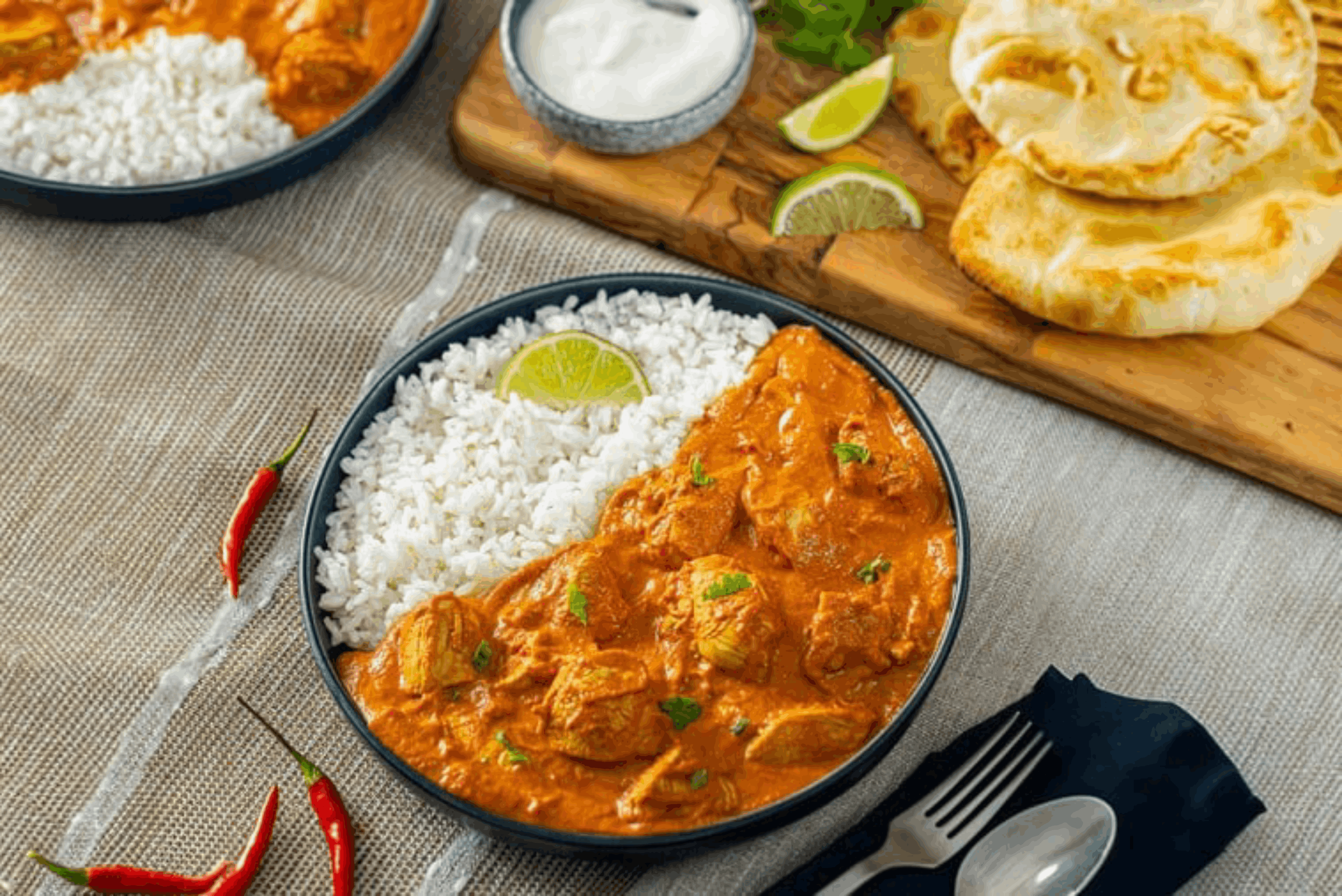 A bowl of Butter Chicken served with white rice, garnished with a lime wedge and cilantro leaves. Accompanied by two pieces of naan bread on a wooden board, a small bowl of raita, and a few red chili peppers. The meal is presented on a light-colored tablecloth with utensils placed beside the bowl.