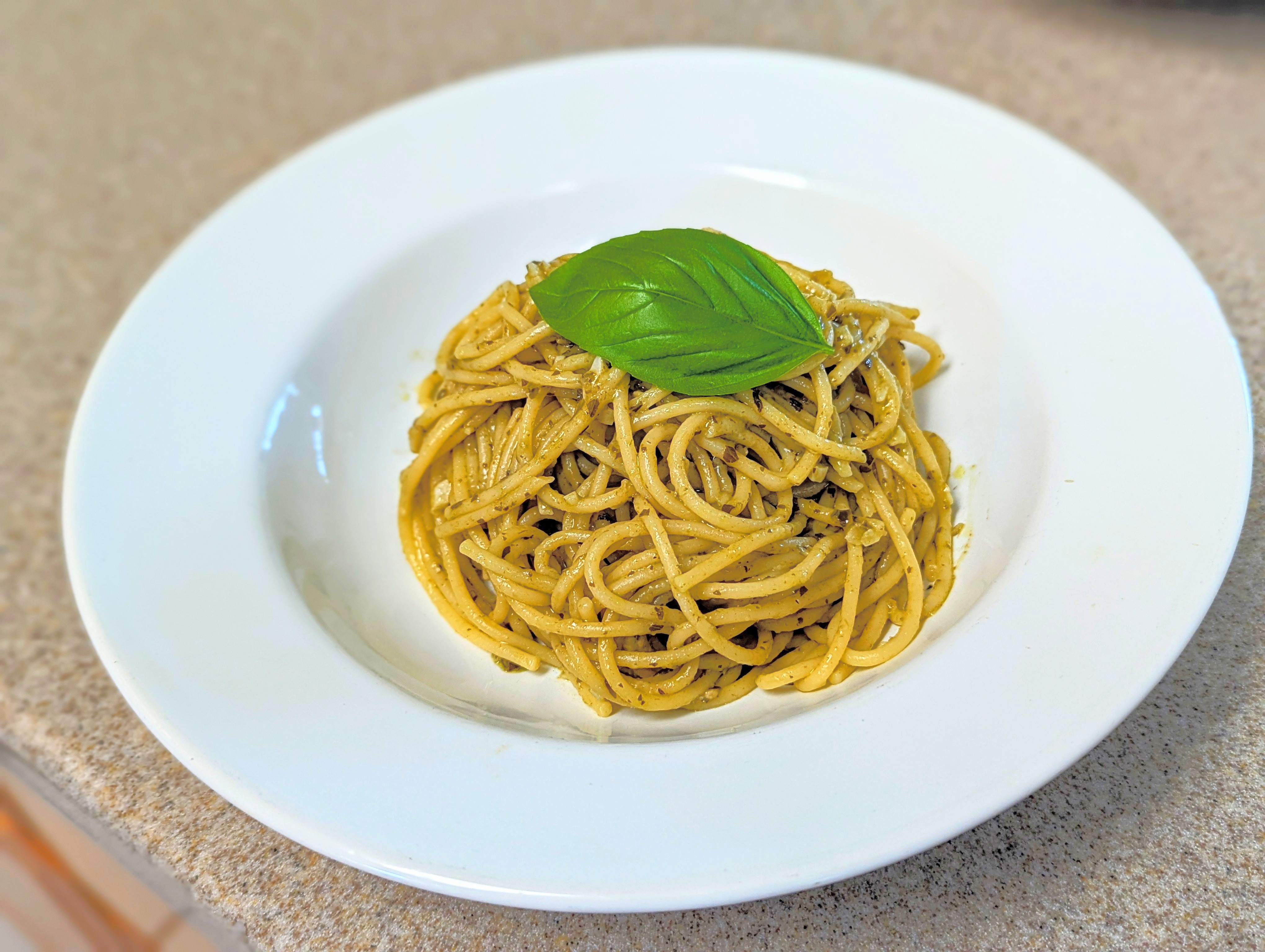 A plate of spaghetti garnished with a single fresh basil leaf on top. The pasta is coated with a pesto sauce, indicated by its greenish tint and visible herb specks. The dish is served on a white ceramic plate, placed on a beige countertop with a speckled pattern