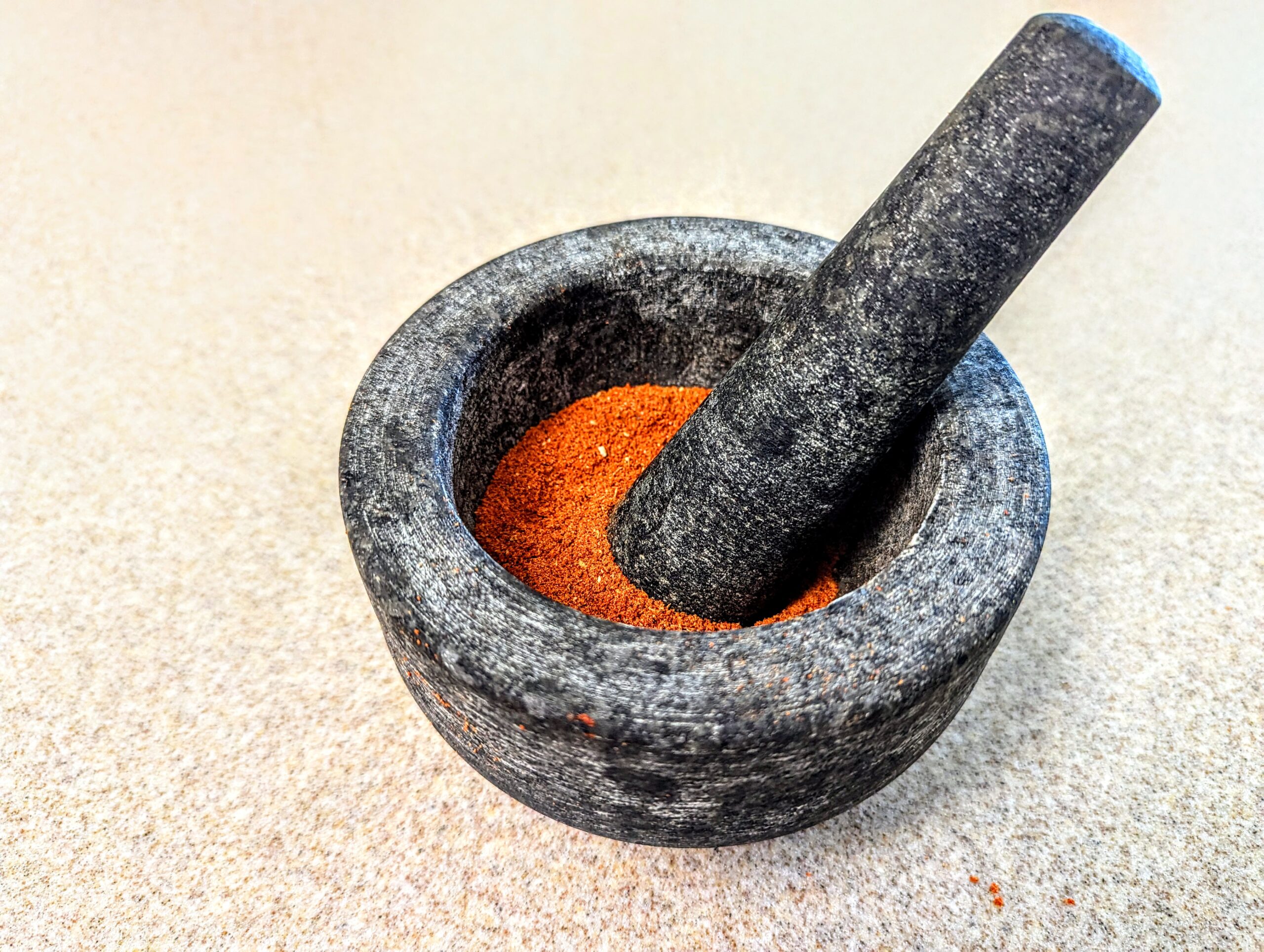 Carne Asada Marinade ingredients in a bowl. A close-up image of a dark grey stone mortar and pestle containing a bright orange powder. The rough, porous texture of the stone contrasts with the fine, smooth quality of the powder. The mortar rests on a beige surface with speckled patterns