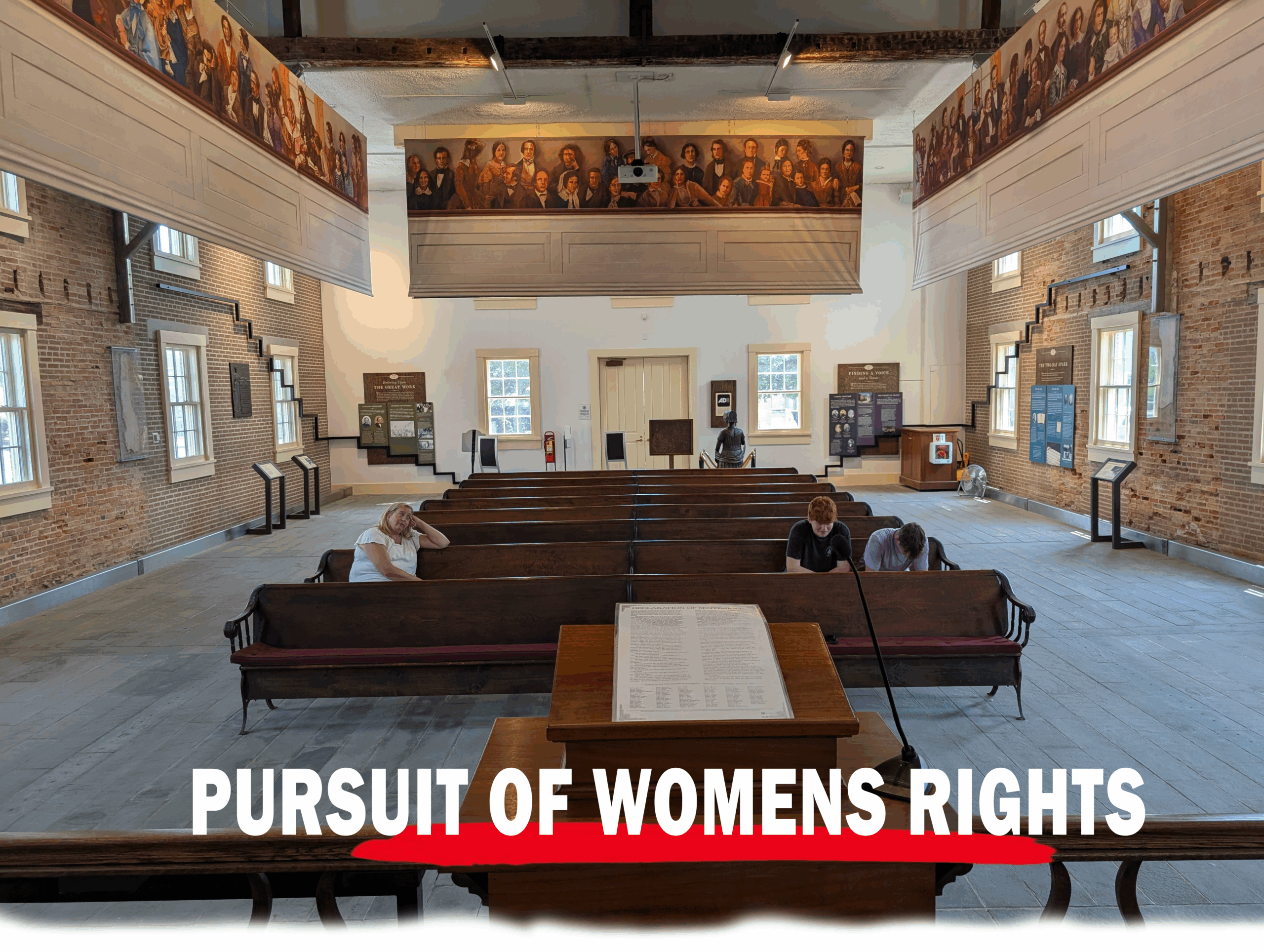 Interior of the historic Wesleyan Chapel at Seneca Falls, with wooden pews, informational displays along brick walls, and a podium at the front. Visitors are quietly reflecting or reading beneath large murals honoring women’s rights pioneers