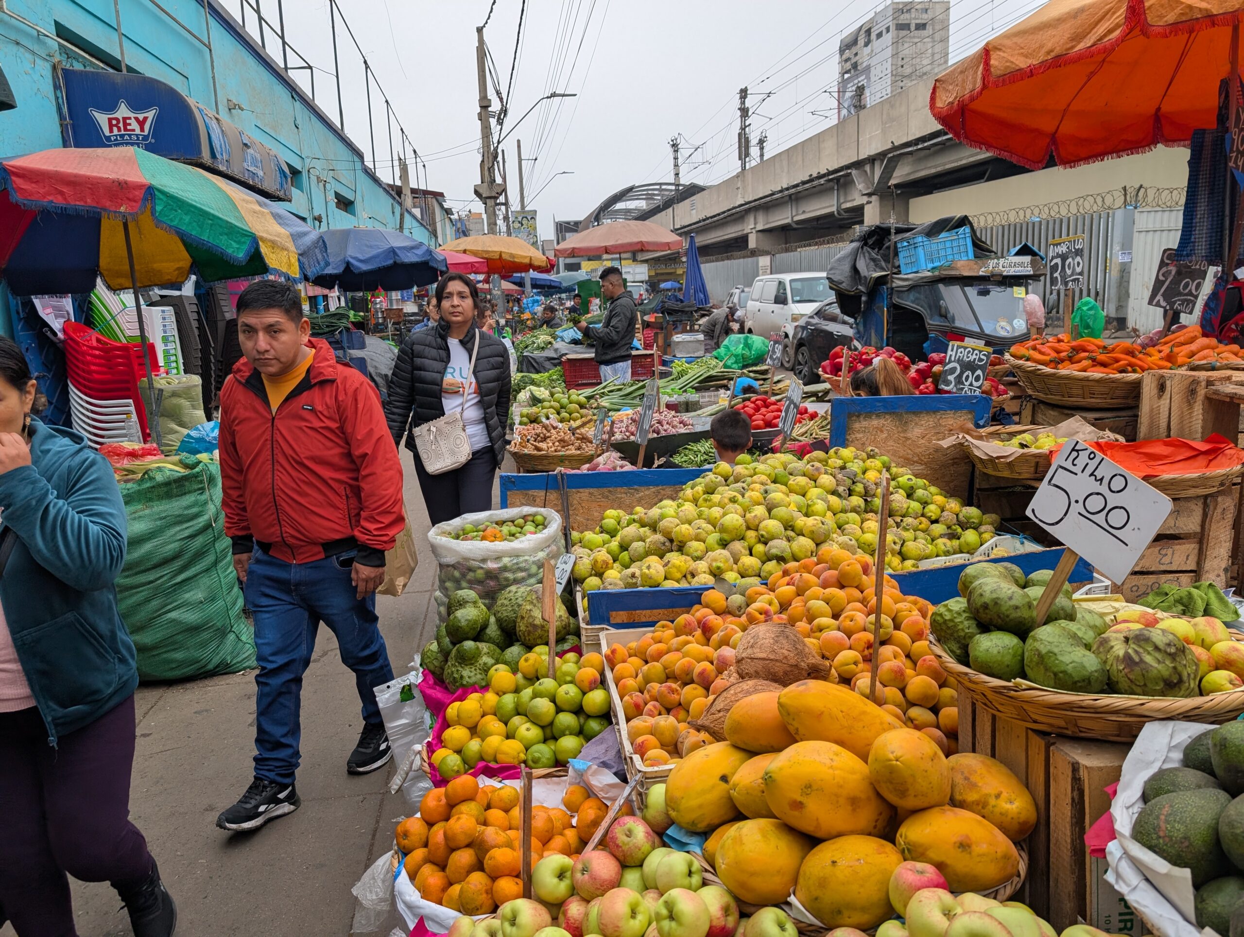 A vibrant outdoor market stall in Lima, Peru, showcasing crates and baskets filled with fresh fruits like papayas, oranges, apples, and pears. Price signs in Spanish hang above the produce, while shoppers move through the busy scene under shade umbrellas.