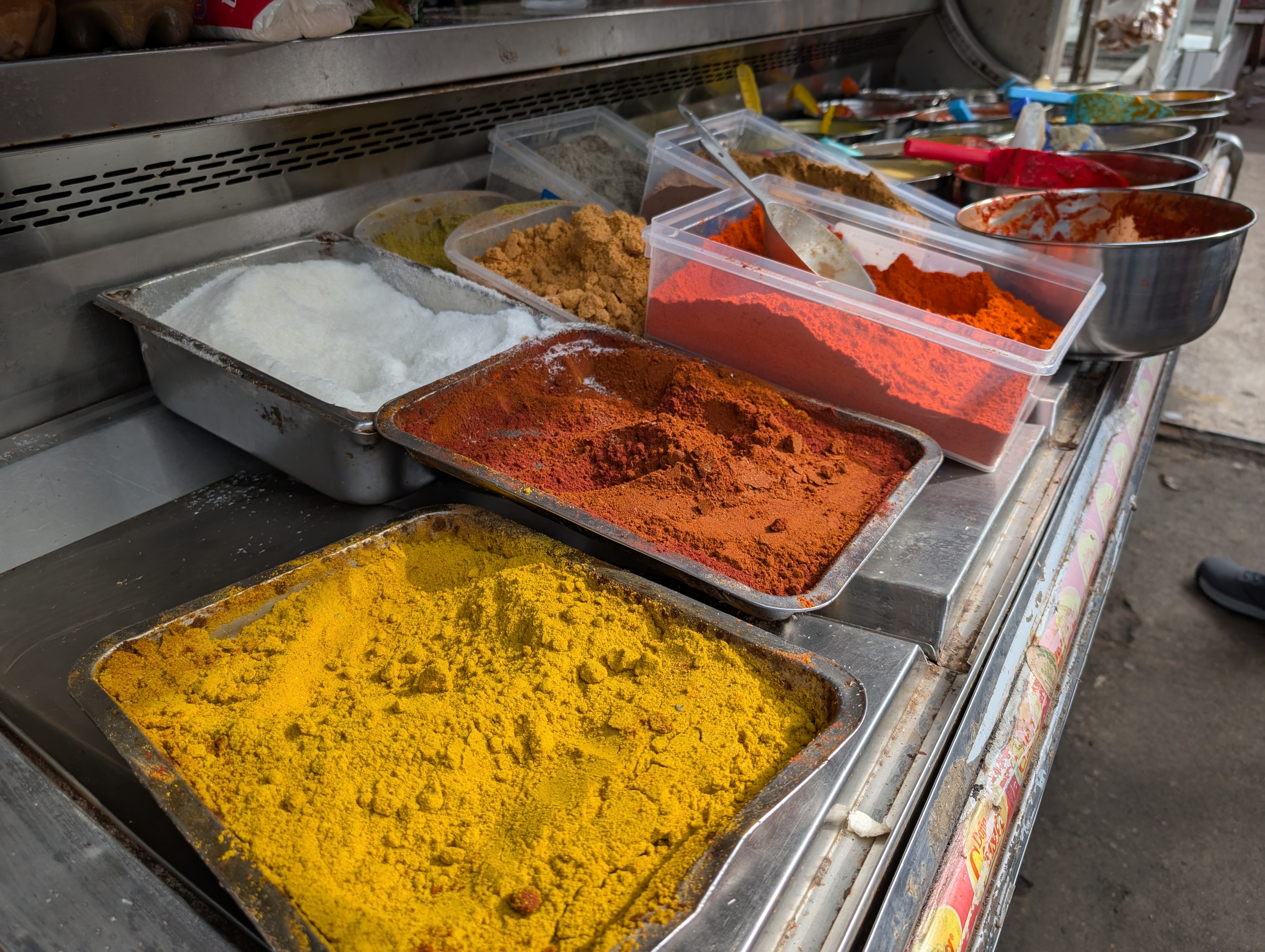A vibrant display of spices at a market stall in Lima, Peru. Metal trays and plastic containers hold colorful powders including red chili, golden turmeric, brown cumin, and white salt. Scoops and spoons rest atop the mounds, capturing the rich culinary diversity of the Mercado Zone.