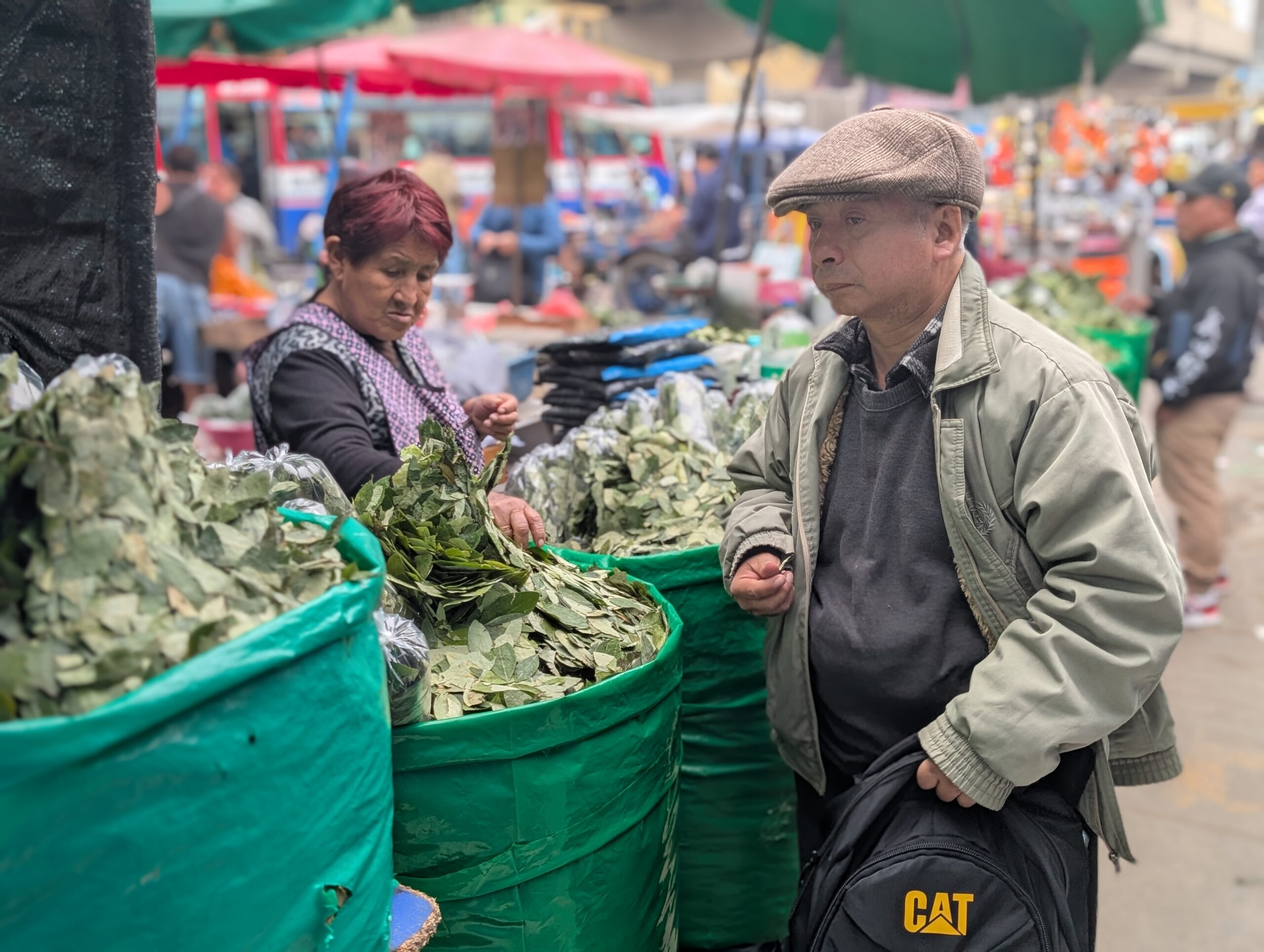A man purchases coca leaves from a street vendor at a market in Lima, Peru. Multiple barrels filled with dried coca leaves line her stall, while surrounding vendors and shoppers create a bustling backdrop