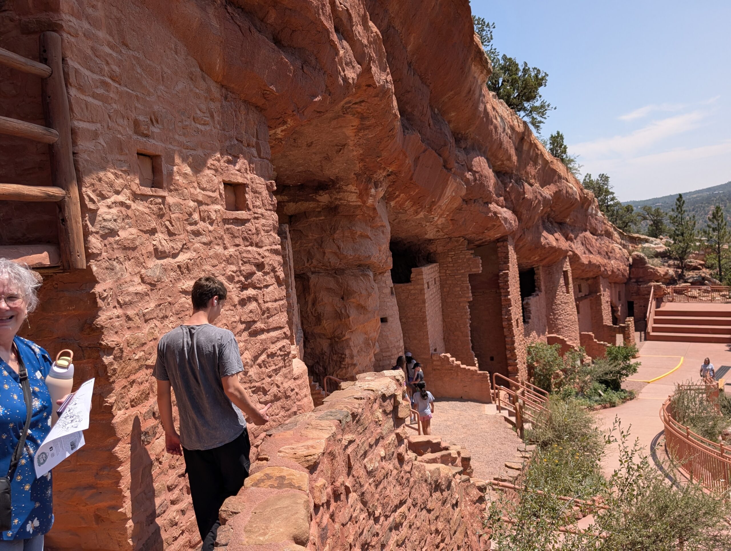 PXL_20260806_193453489 Visitors walking through the Manitou Cliff Dwellings beneath a red sandstone overhang in Colorado